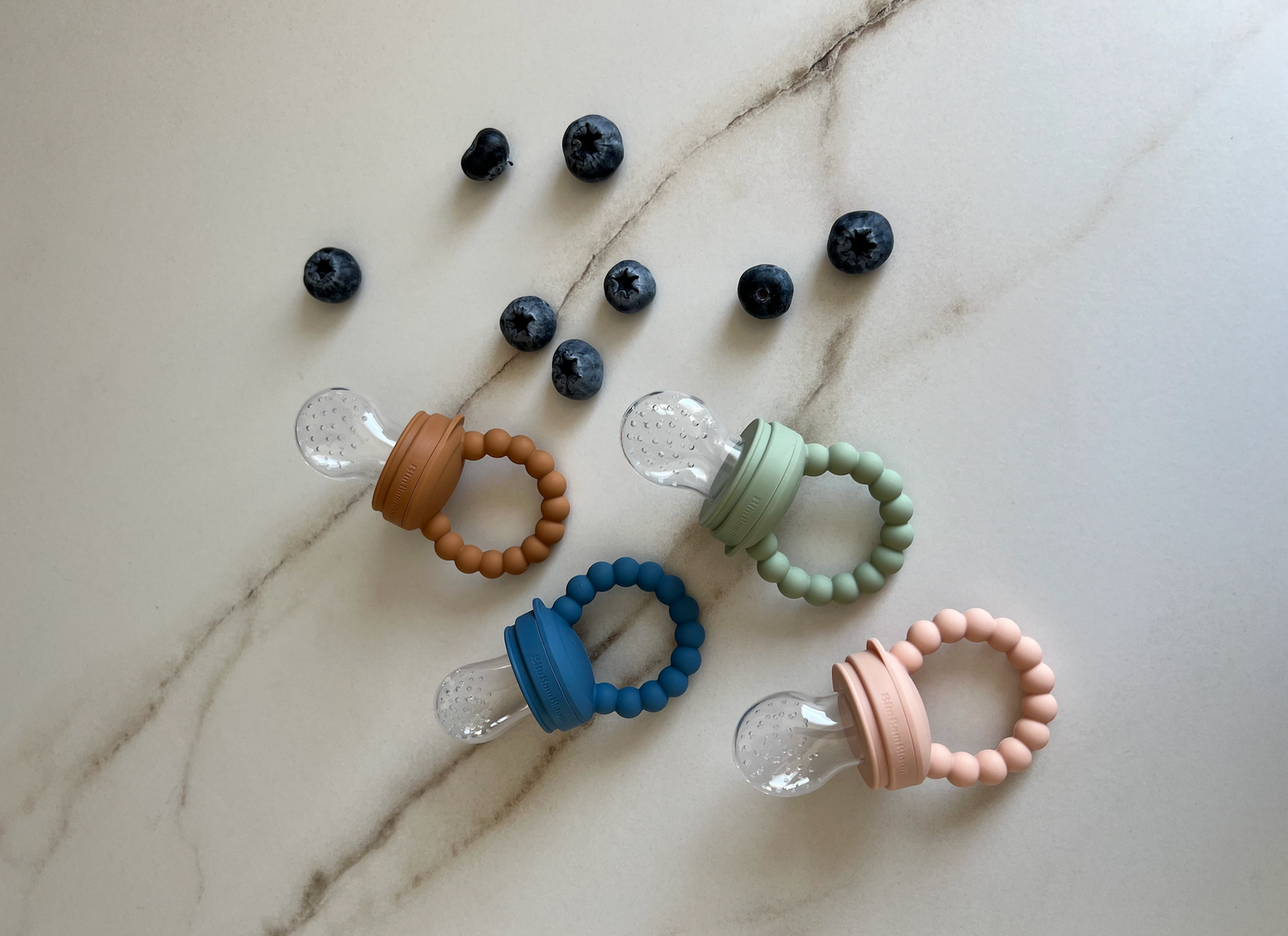 Tetine grignoteuse a fruit pour bebe en couleur bleu, vert, rose et camel sur un plan de travail en marbre blanc et des myrtilles, par Bimbambloom for Cool Families