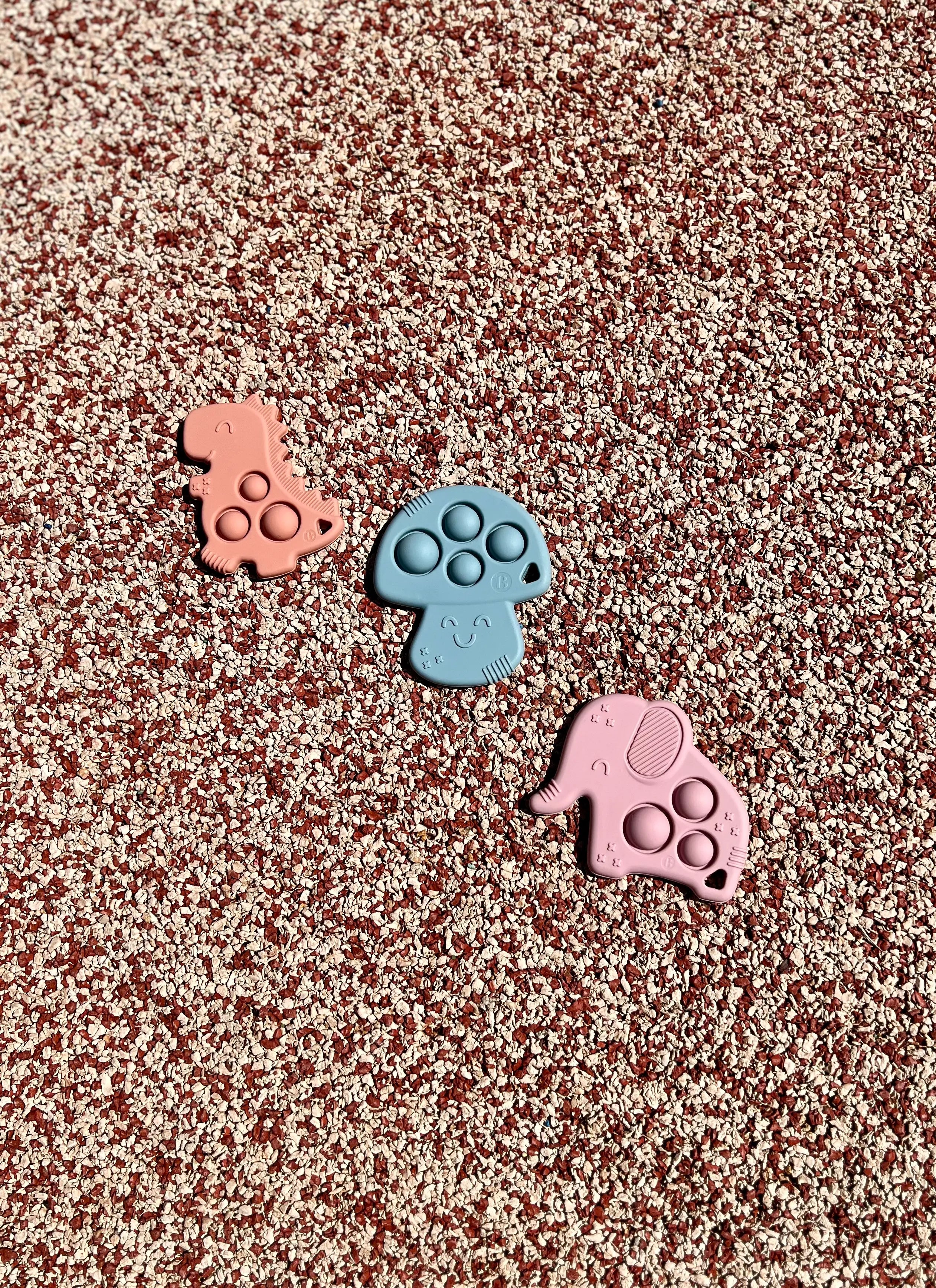 Anneau de dentition Pop-It de Bim Bam Bloom sur un fond pastel, offrant un design ludique et coloré pour soulager les gencives de bébé. Un elephant rose, un champignon bleu et un dinosaure rose.