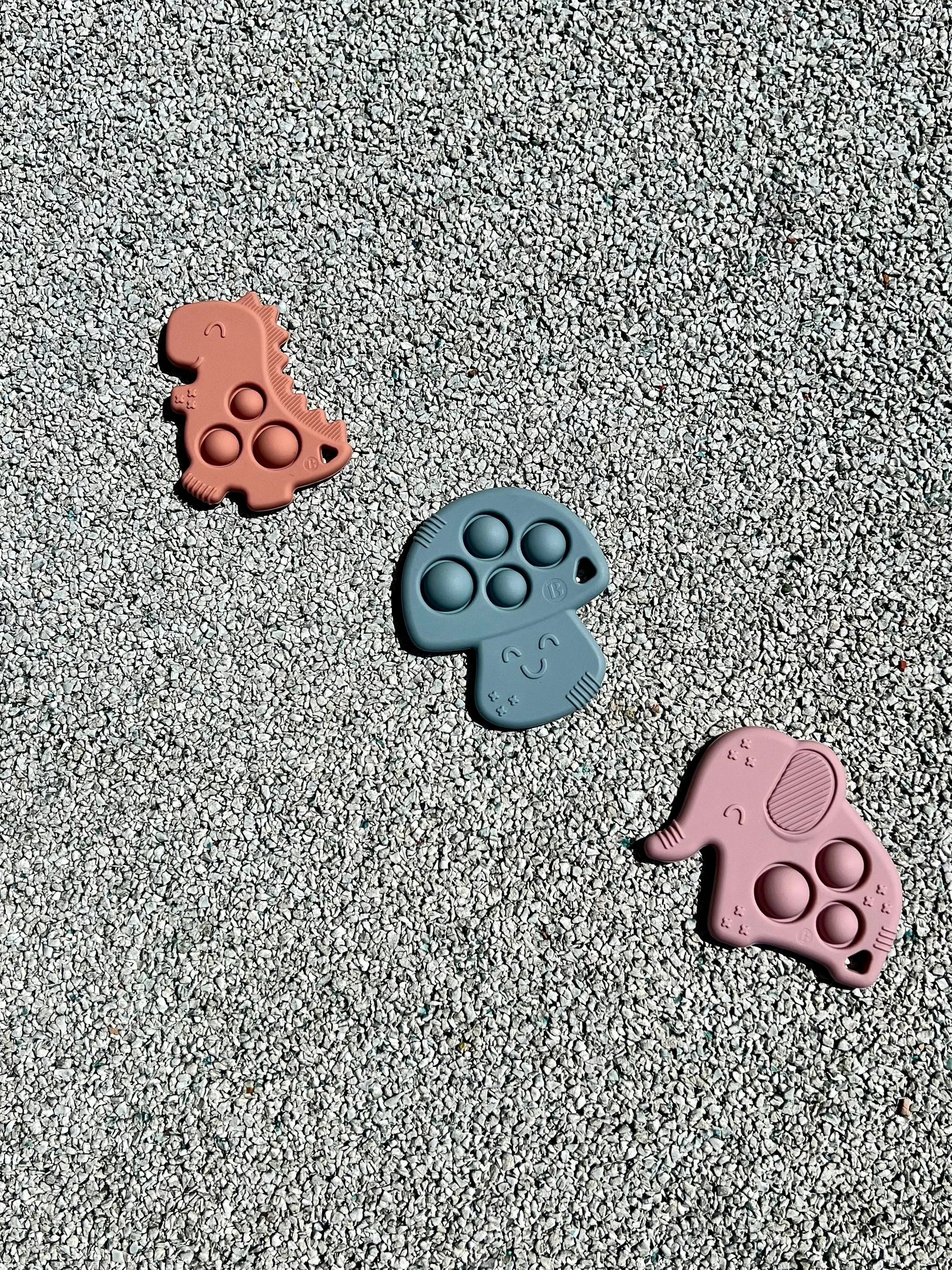 Anneau de dentition Pop-It de Bim Bam Bloom sur un fond pastel, offrant un design ludique et coloré pour soulager les gencives de bébé. Un elephant rose, un champignon bleu et un dinosaure rose.