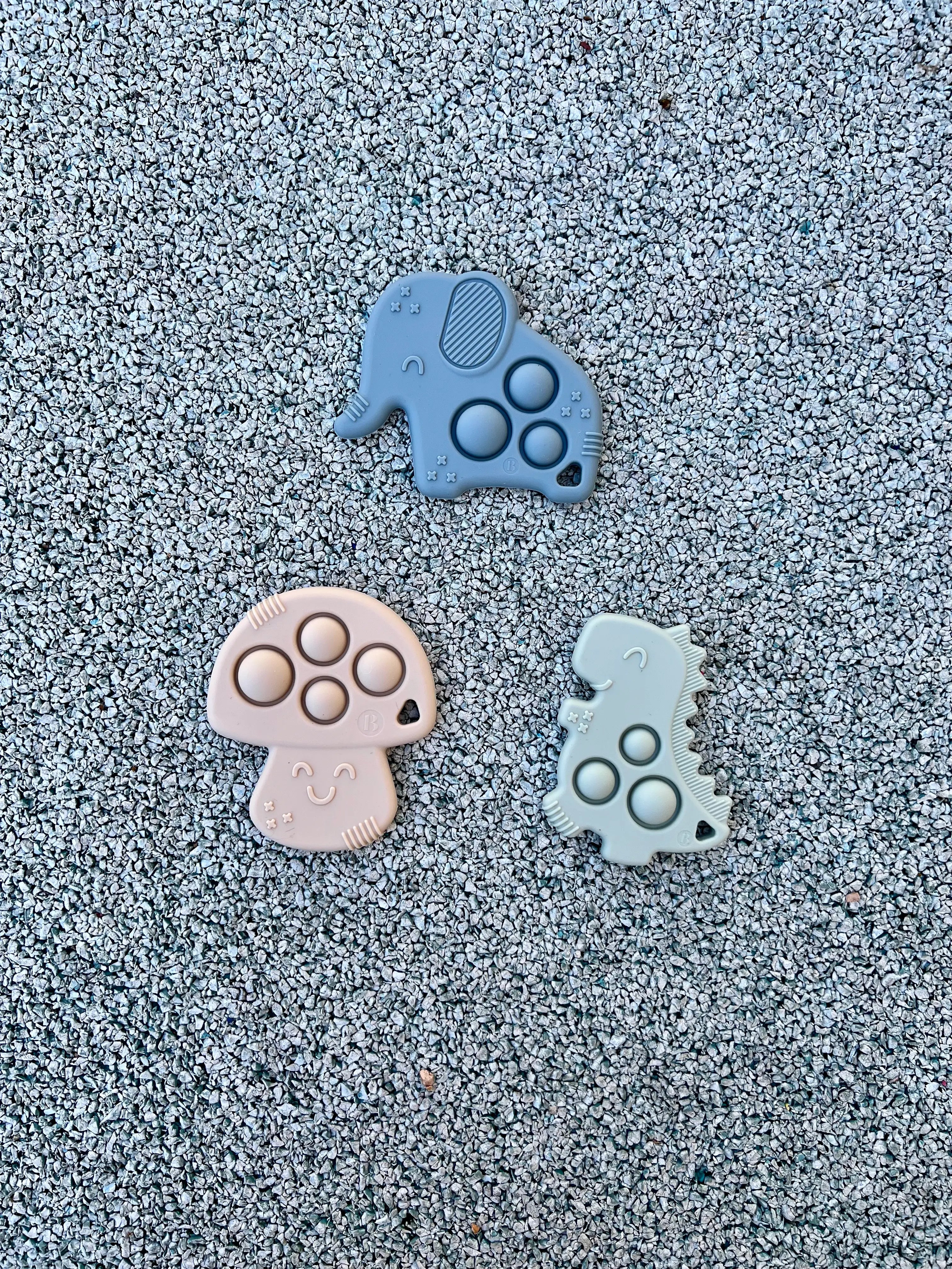 Anneau de dentition Pop-It de Bim Bam Bloom sur un fond pastel, offrant un design ludique et coloré pour soulager les gencives de bébé. Un elephant bleu, un champignon beige et un dinosaure vert.