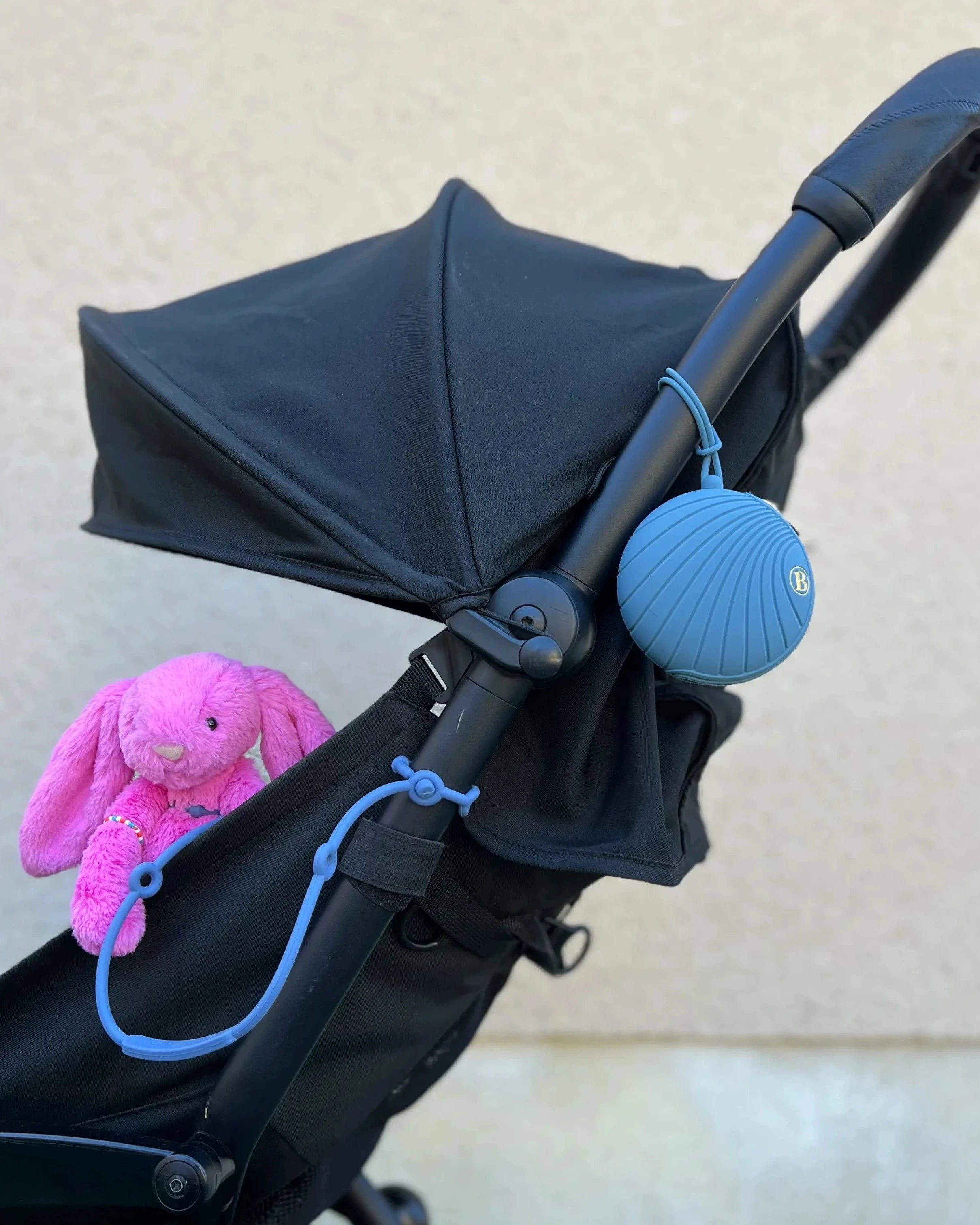 Boîte à tétine bleue Bim Bam Bloom attachée à la poussette avec un attache-doudou Lovie Link bleu, pour ne plus jamais perdre le précieux doudou de votre bébé