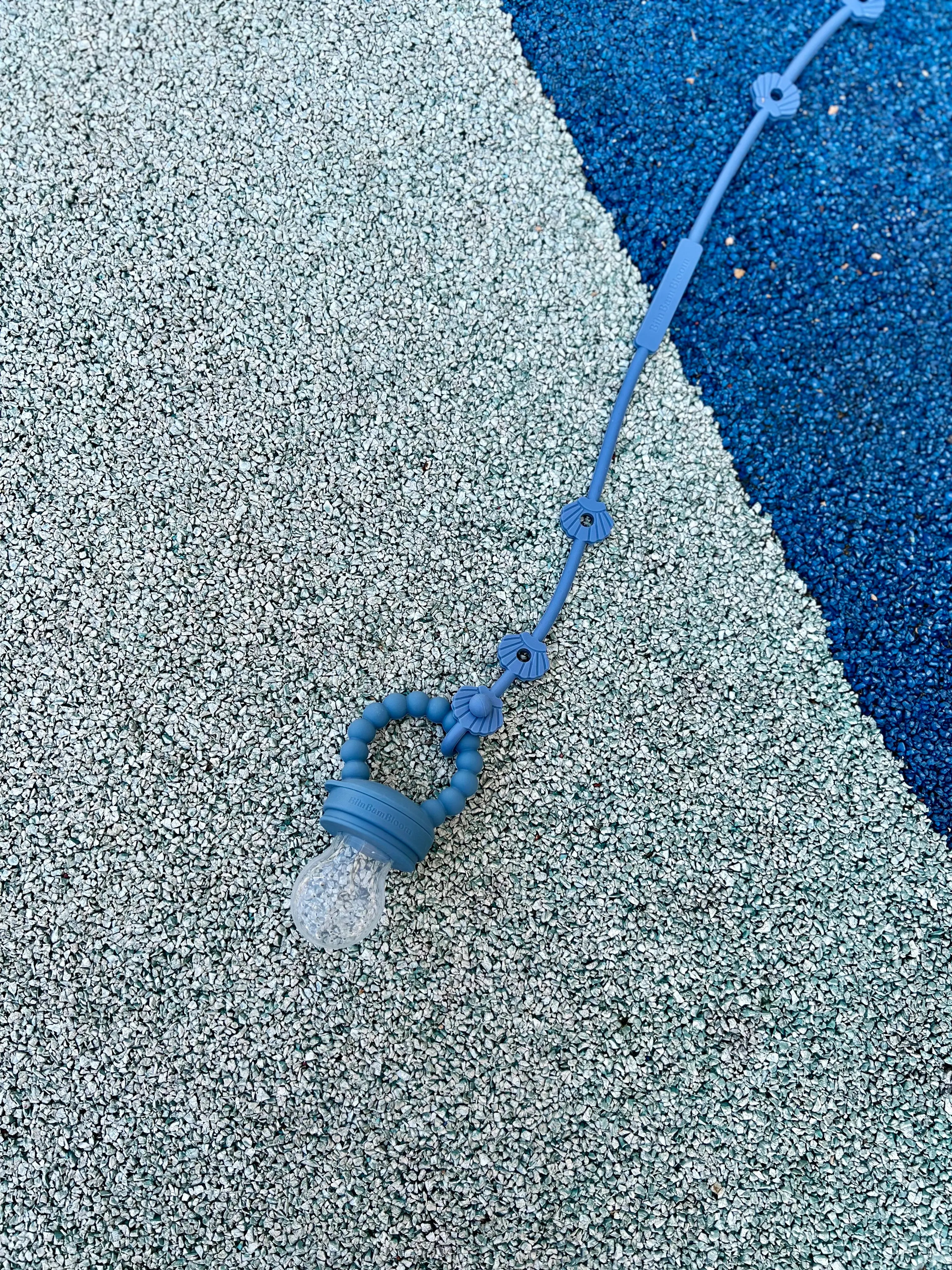 Tetine grignoteuse, gourmande, a fruit pour bebe en silicone Bleu Bim Bam Bloom avec son attache pour pouvoir l'accrocher a la poussette et a la chaise haute.