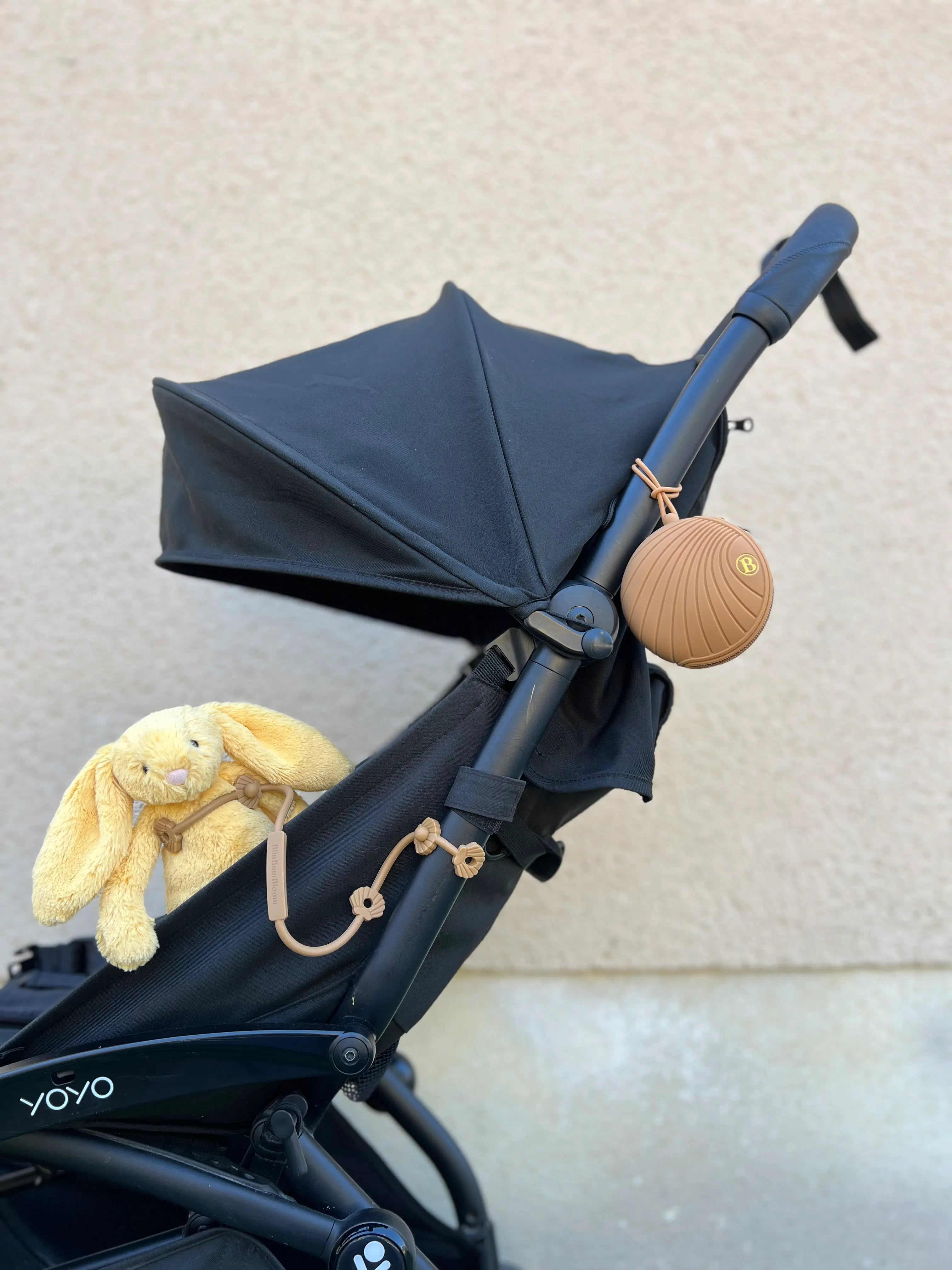 Laisse à doudou Lovie Link® Shell camel dune dorée Bim Bam Bloom attachée à une poussette noire, avec une boîte à tétine Bim Bam Bloom, pour des sorties sereines et organisées.