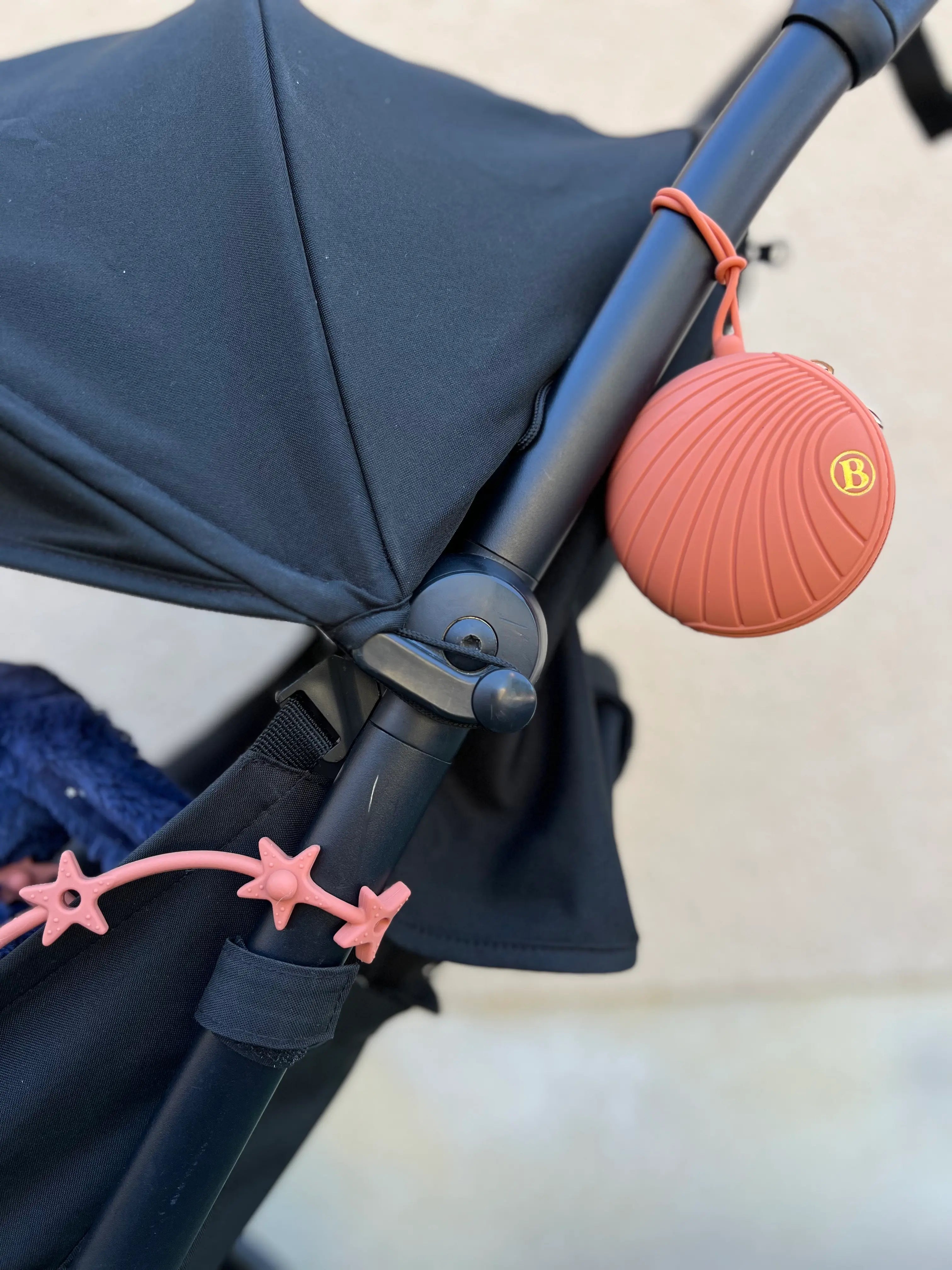 Laisse à doudou Lovie Link® Terracotta fixée à une poussette, garantissant des sorties sereines et organisées pour les parents. Boite à tétine en silicone Bim Bam Bloom Terracotta fixée à la poussette.