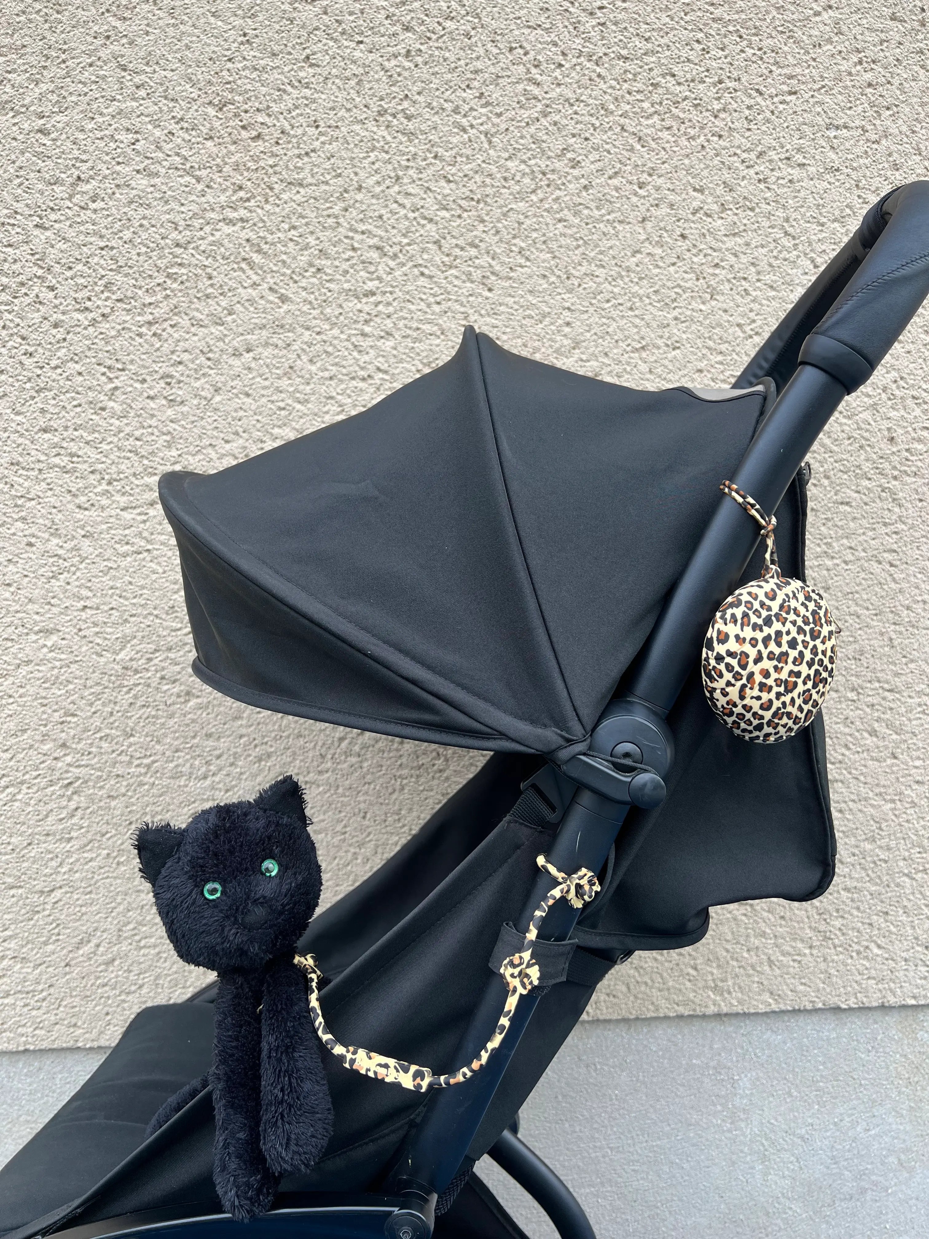 Attache doudou pour poussette Lovie Link® Shell imprimé léopard attachée à une poussette noire, accompagnée d'une boîte à tétine Bim Bam Bloom, pour des sorties élégantes et organisées.