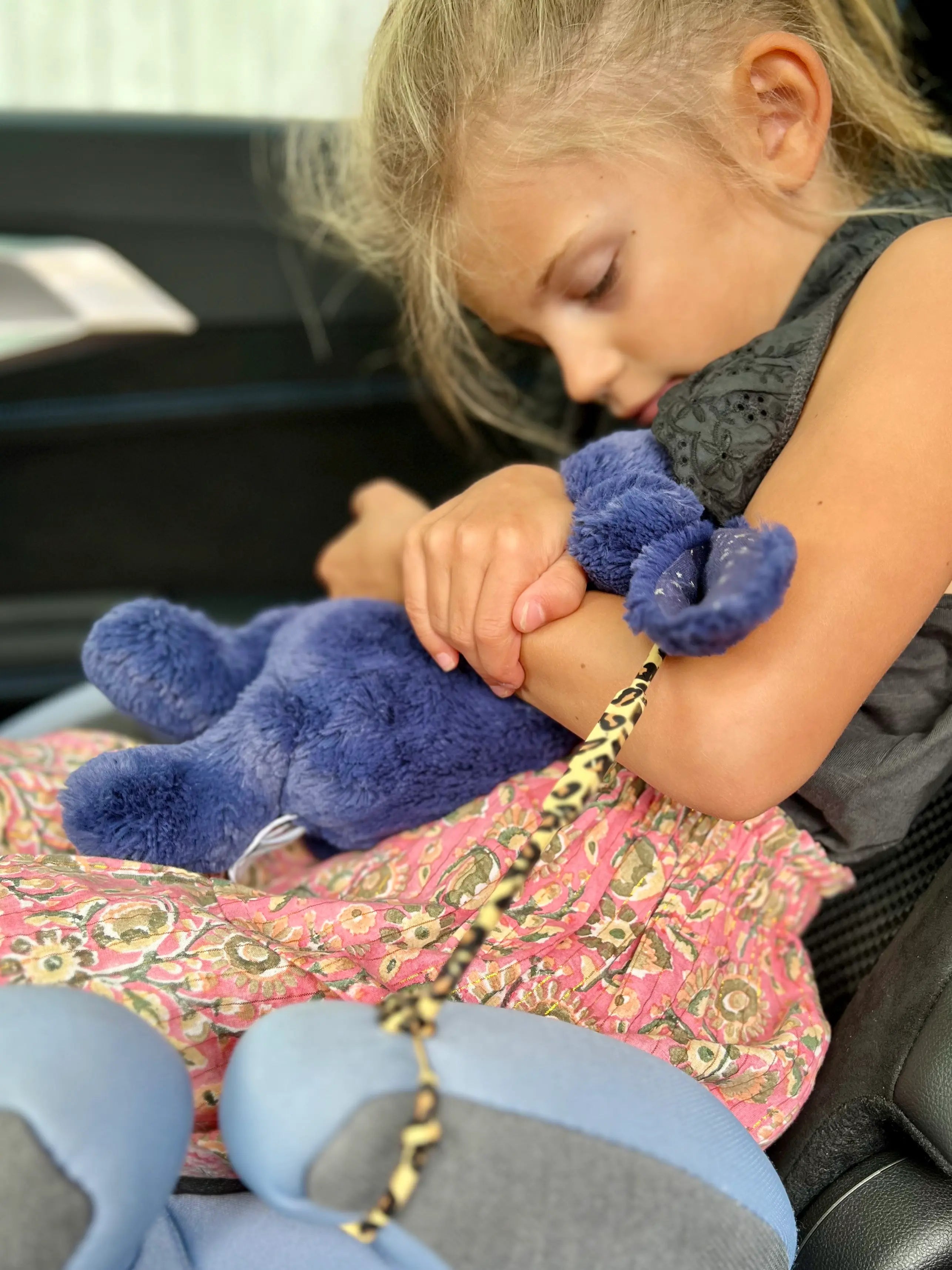 attache doudou Lovie Link® Shell en imprimé léopard, attaché au siege auto de la voiture accroché à un doudou lapin dans les bras d'une petite fille par Bim Bam Bloom. Accessoire essentiel pour garder le doudou de bébé toujours à portée de main.