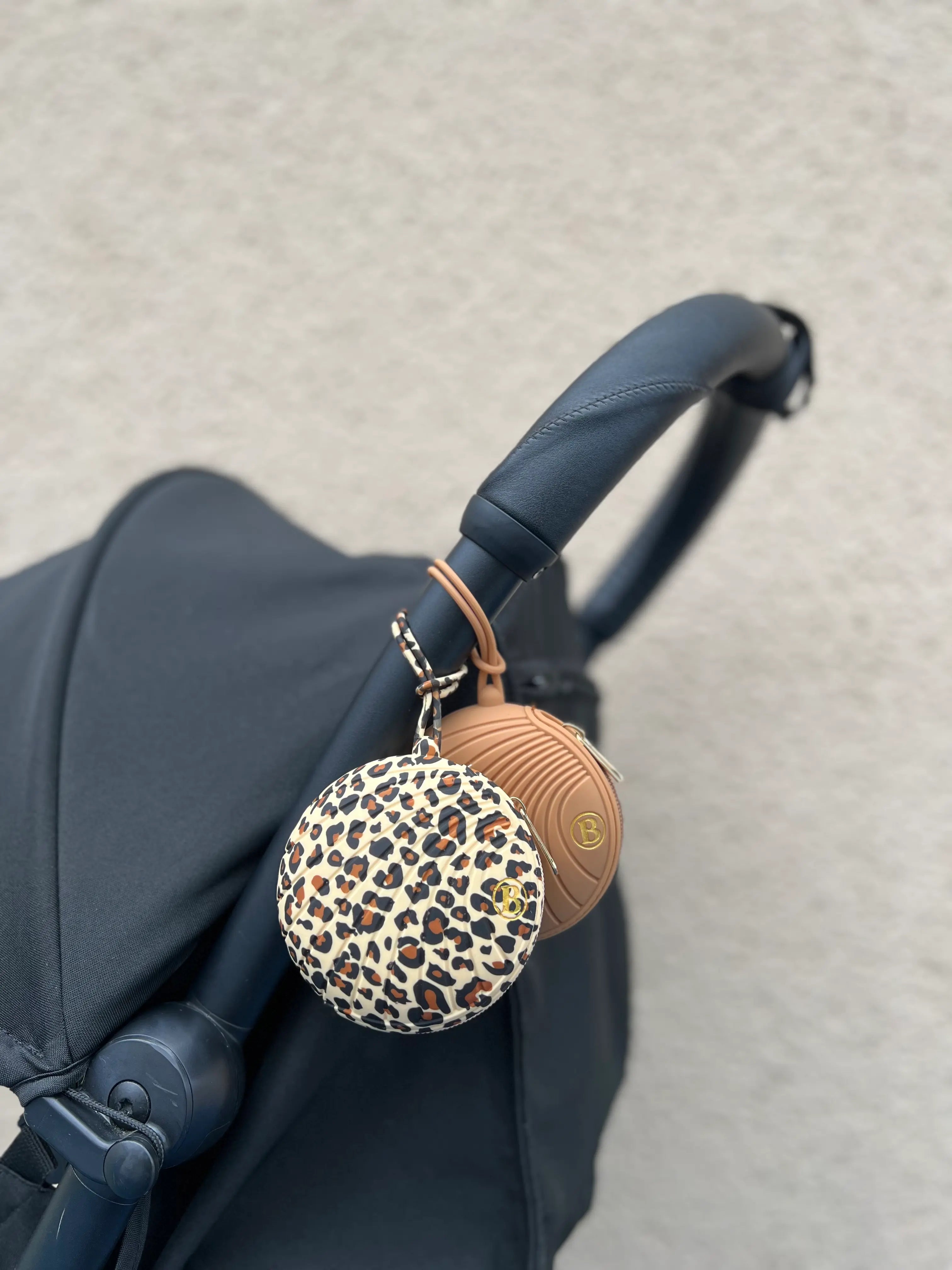Boîtes à tétine en silicone léopard et terracotta attachées à une poussette. Etuis à sucettes Bim Bam Bloom en silicone et à zip.