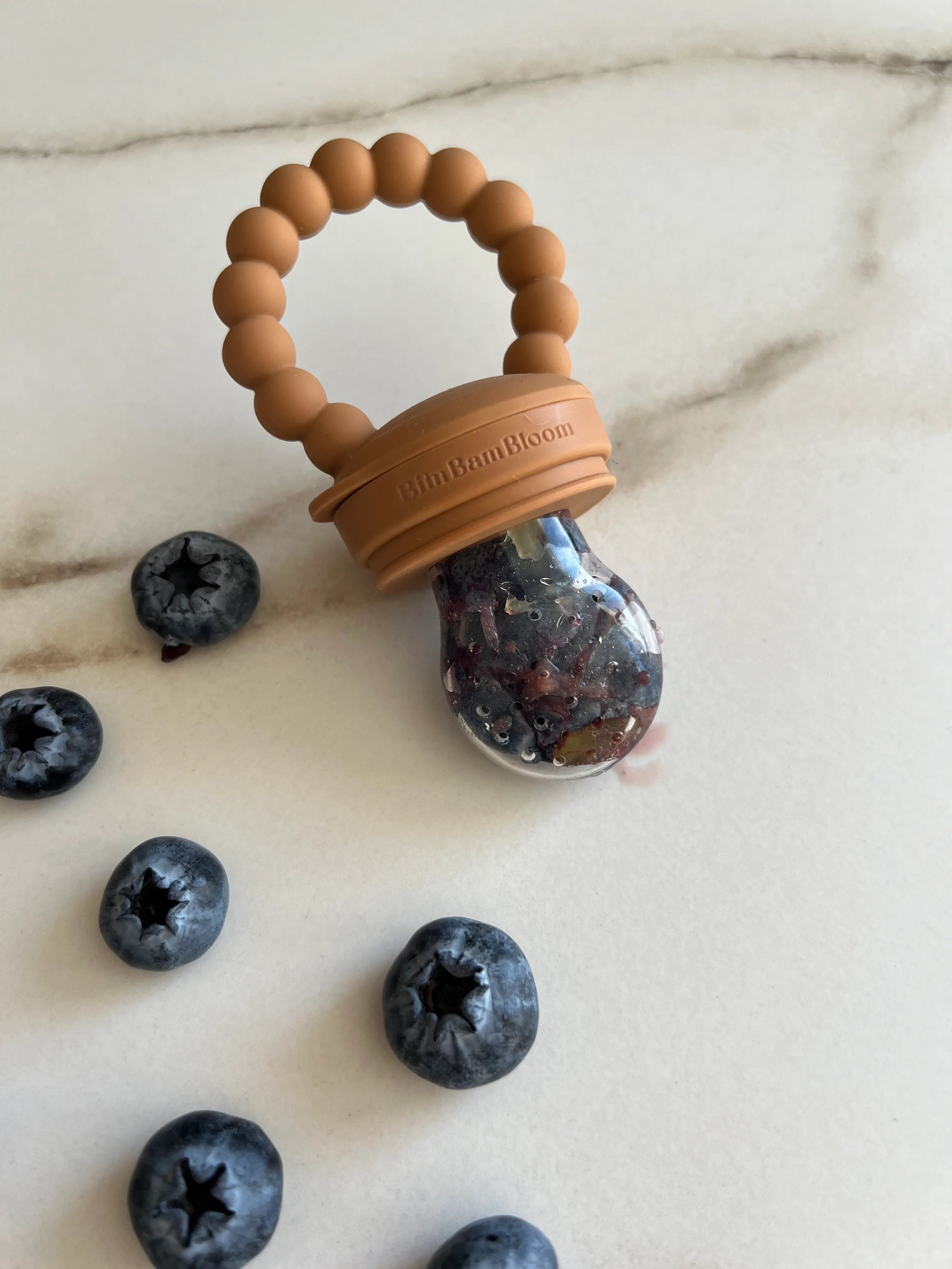 Gros plan sur la tétine grignoteuse Yummy Tot® en Camel, avec des myrtilles, idéale pour faciliter l'apprentissage de la diversification alimentaire avec un design sûr et amusant pour bébé.
