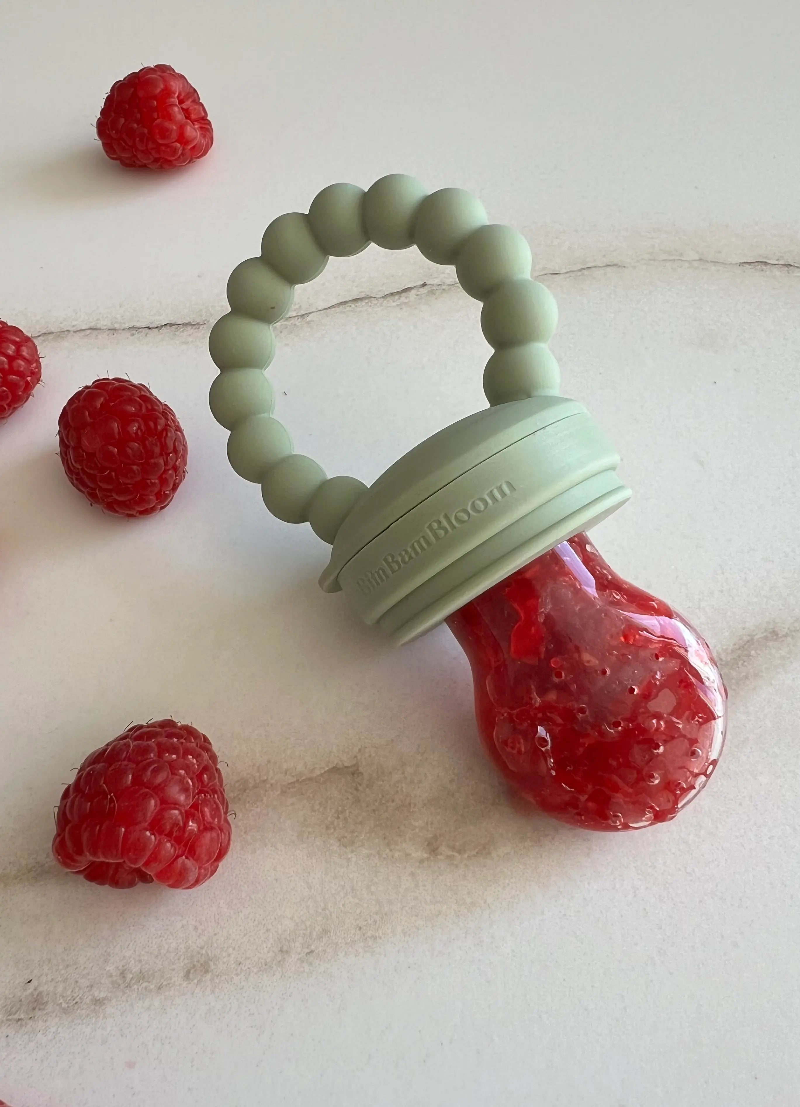 Tétine grignoteuse en silicone vert Bim Bam Bloom. Gros plan sur la tétine remplie de framboise et son jus se formant sur la tétine gourmande.