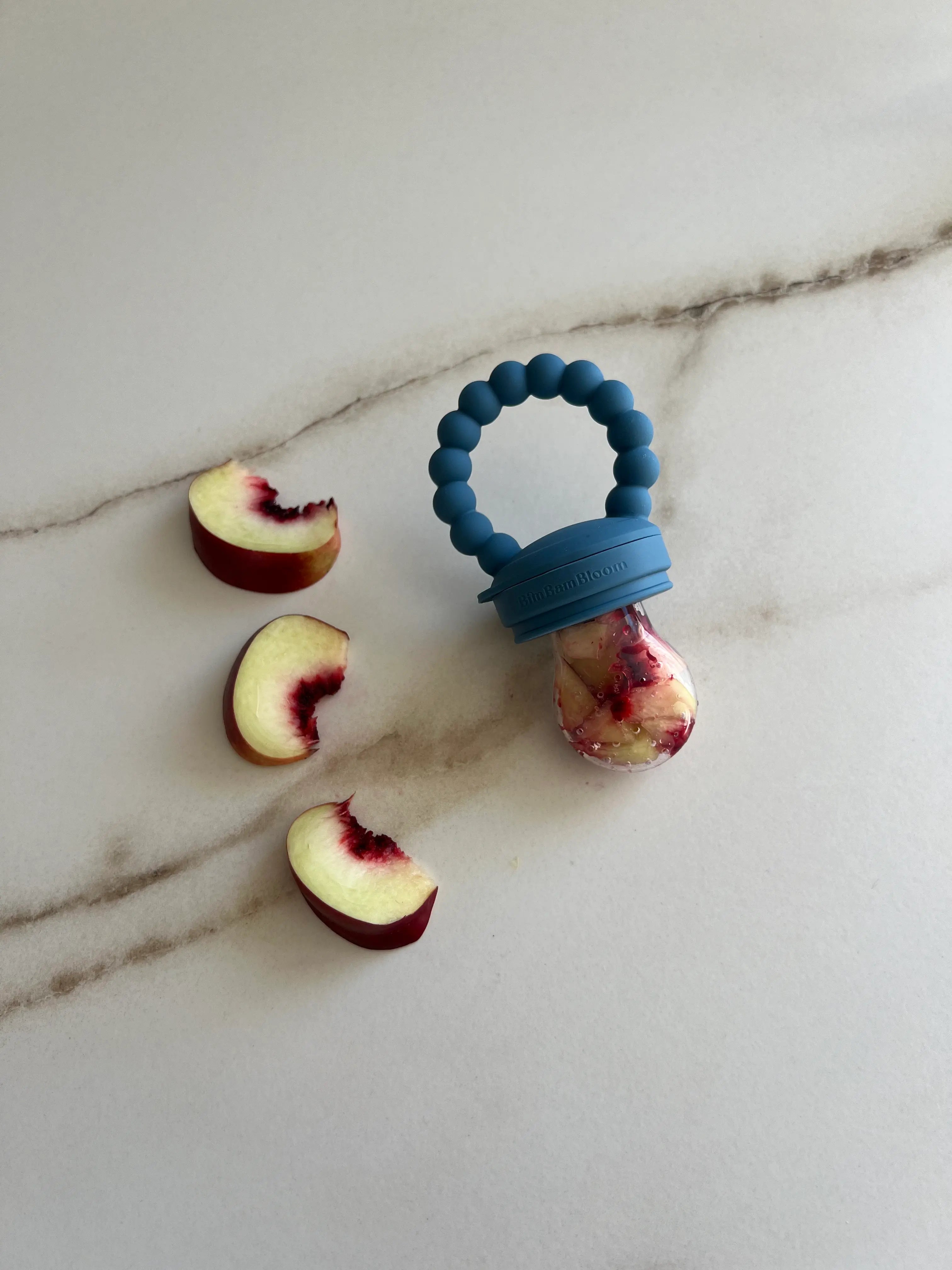 Tetine grignoteuse, gourmande, a fruit pour bebe en silicone Bleu Bim Bam Bloom remplie de morceaux de nectarines et quartier de nectarines autour.