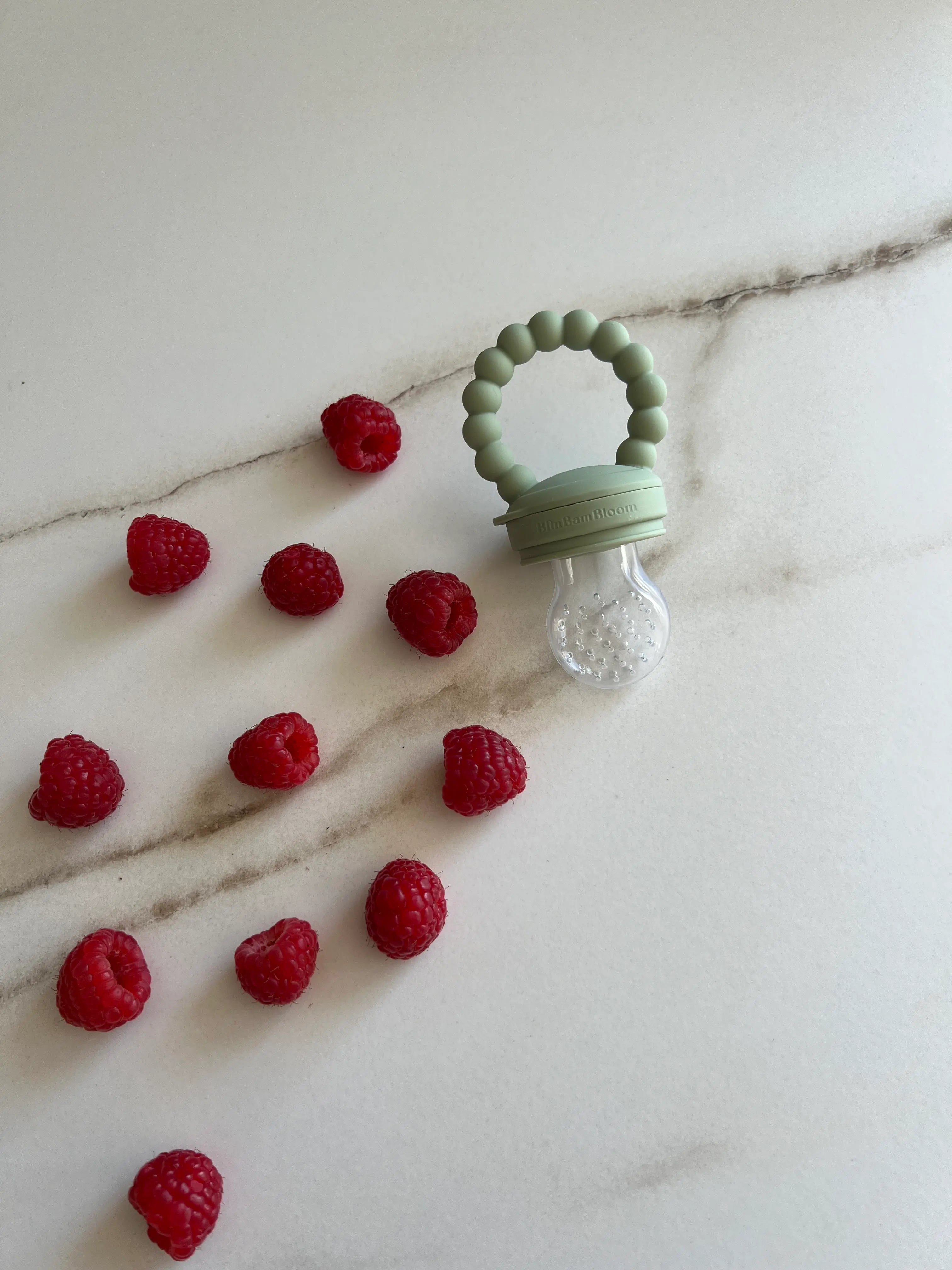 Tétine grignoteuse en silicone vert Bim Bam Bloom avec des framboises sur le plan de travail.