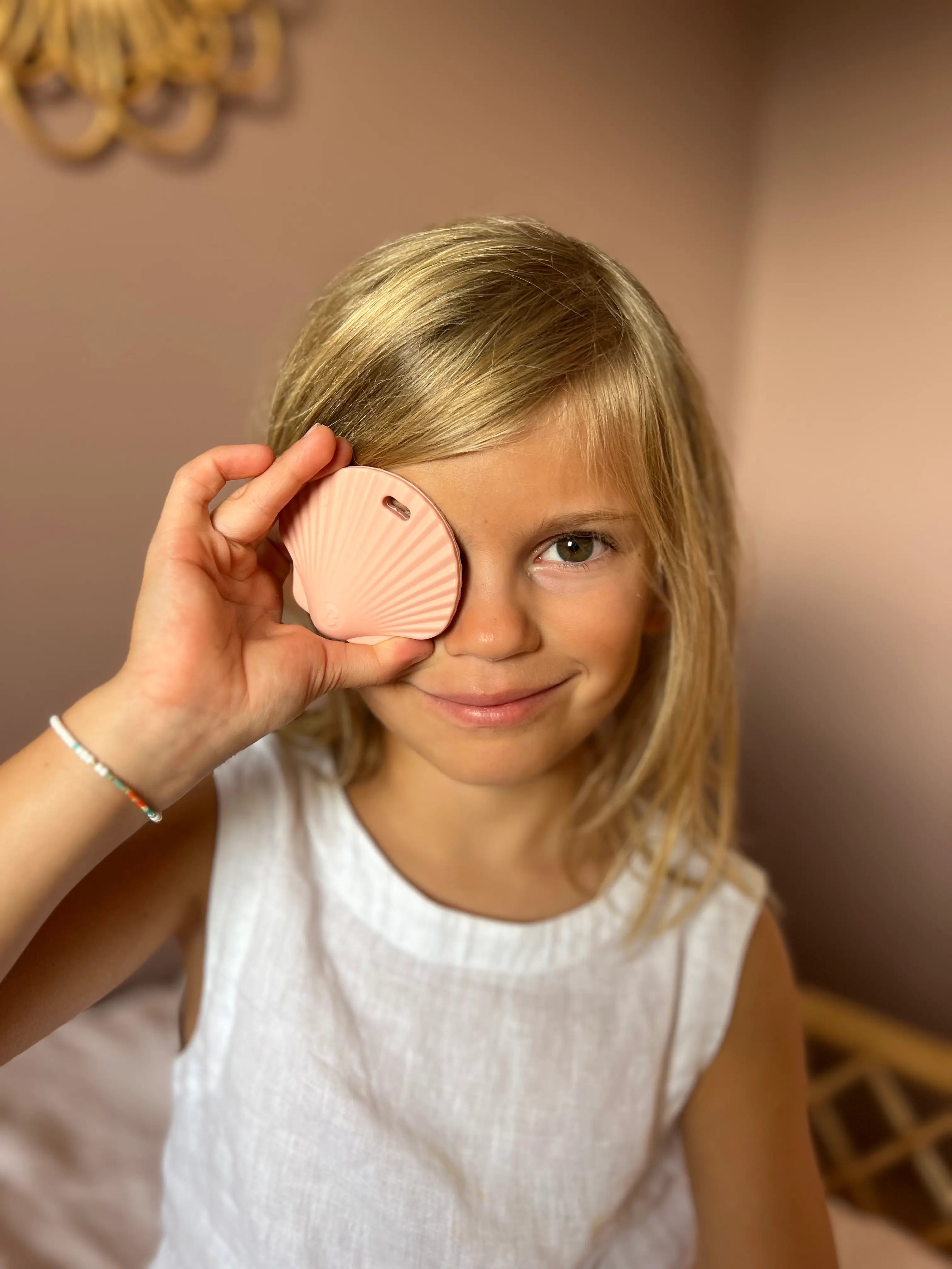 Petite fille tenant l'anneau de dentition Shell Rose Guimauve Fondante devant son oeil, un jouet de dentition doux et réconfortant pour soulager les gencives de bébé par Bim Bam Bloom