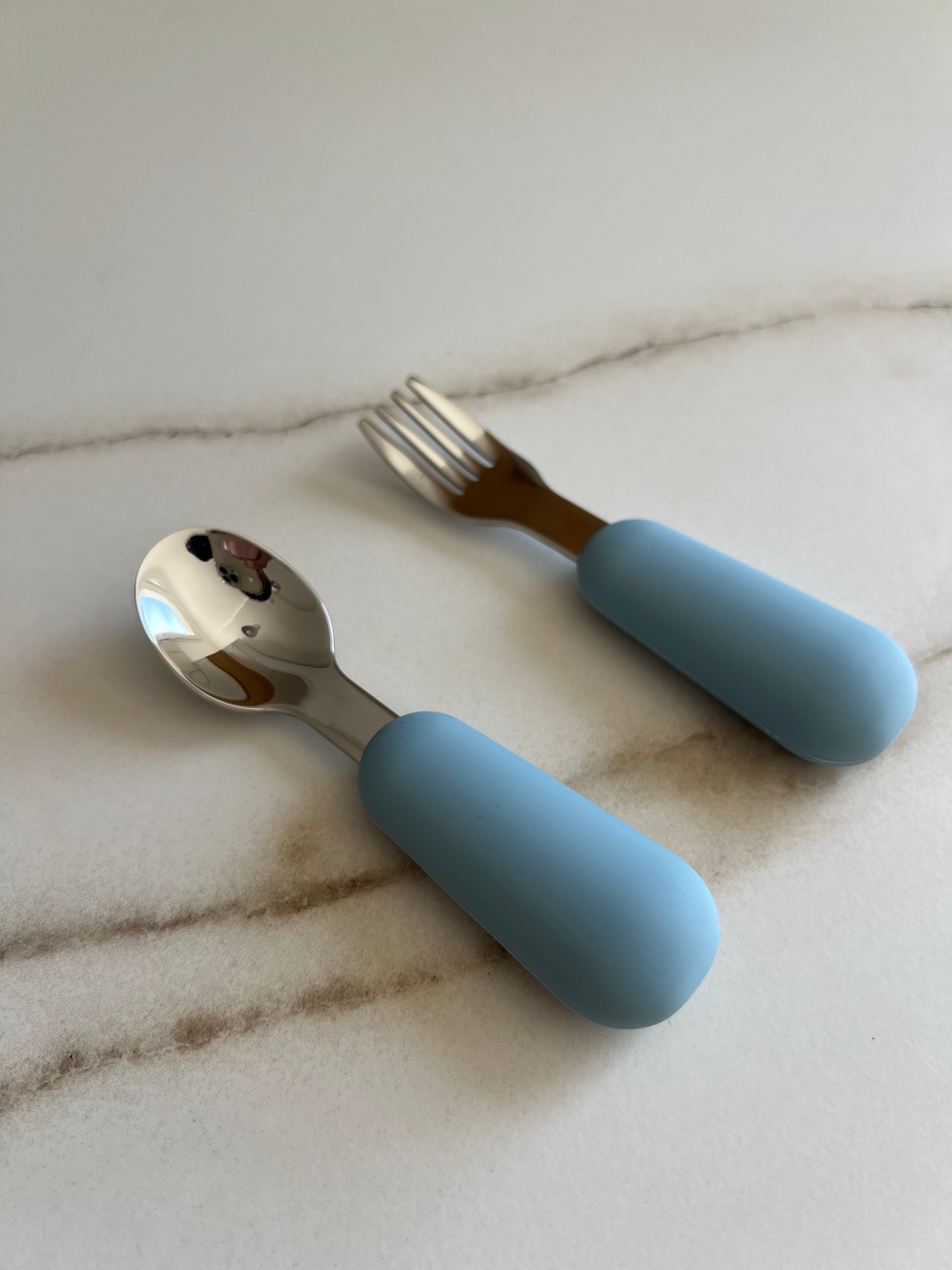 Set de couverts bébé bleu en inox et silicone posé sur une table cuillère et fourchette adaptées aux petites mains